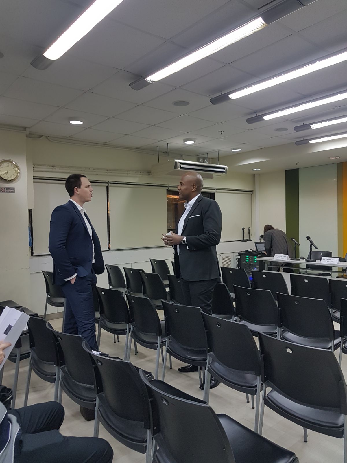 Two men in suits talk in an empty conference room with rows of chairs.