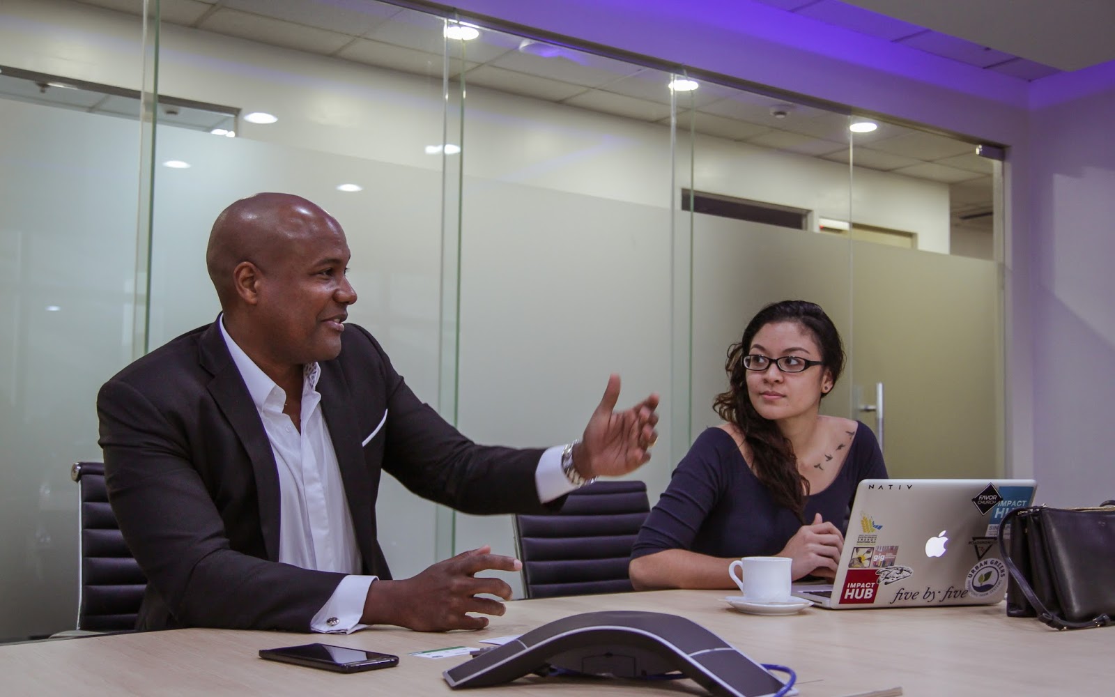 Man in suit talking to woman at laptop in modern office meeting room.