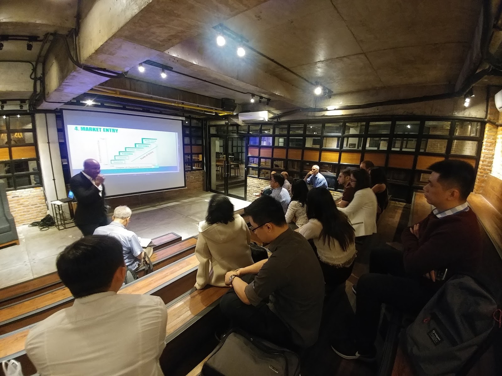 Speaker presenting "4. Market Entry" slide to an audience seated on tiered wooden benches.