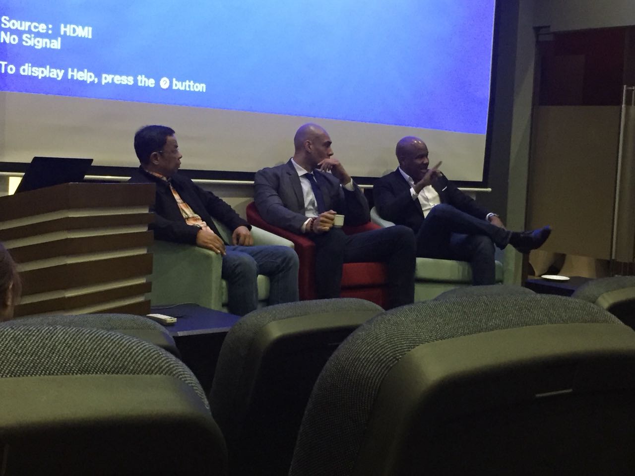 Three men sit on chairs in front of a large screen displaying "Source: HDMI No Signal".