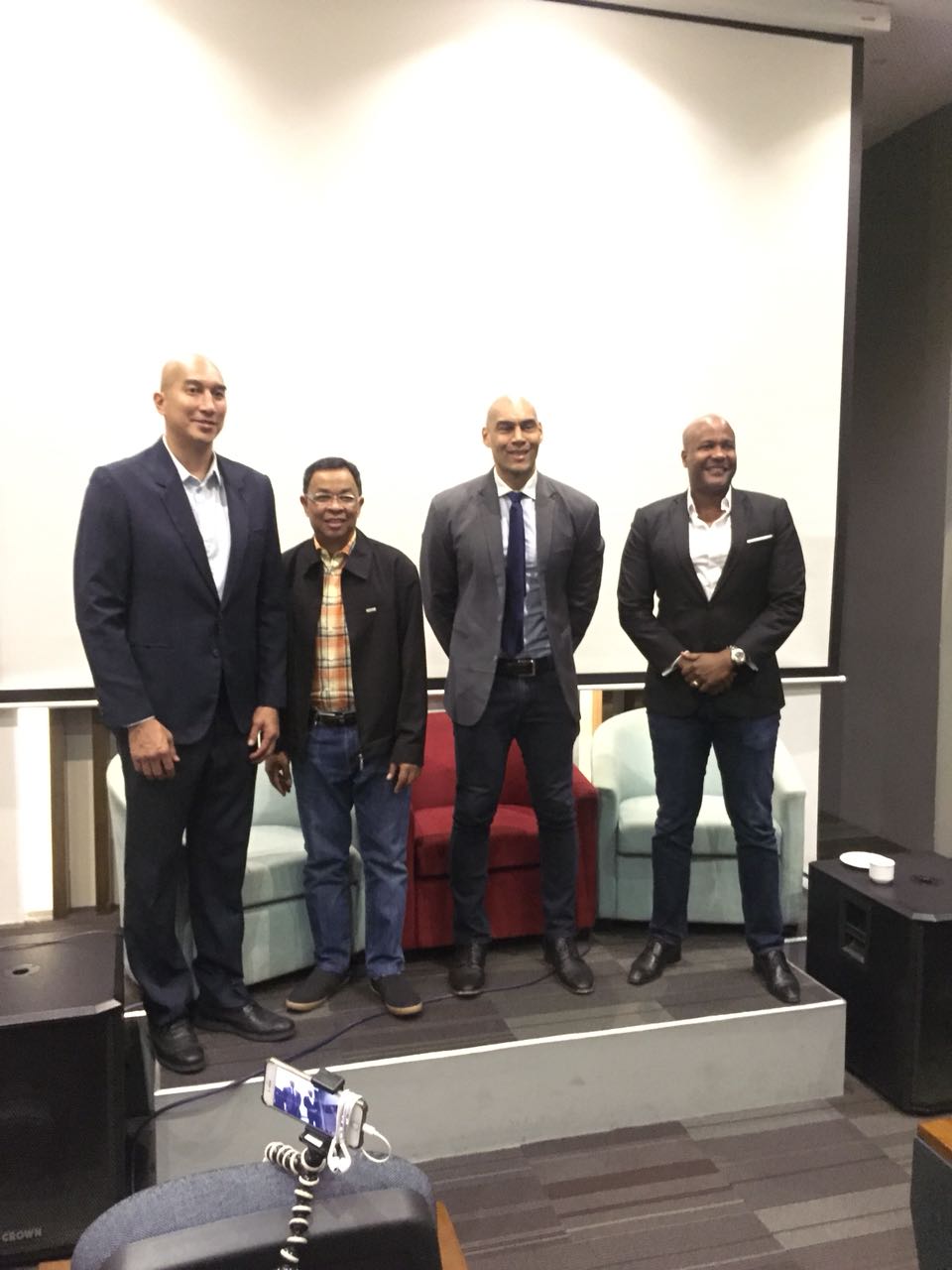 Four men stand on a stage in front of a projection screen. Two chairs are visible behind them.