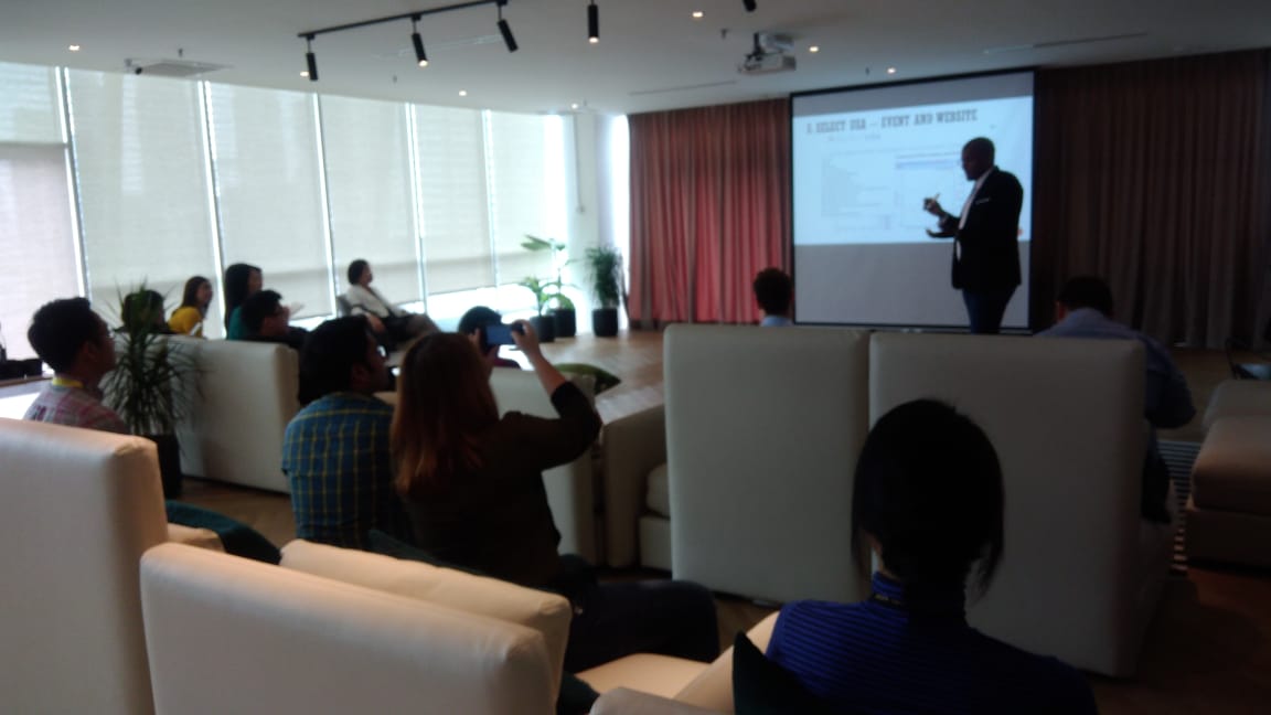 Speaker presents to an audience in a modern conference room with a projector screen.