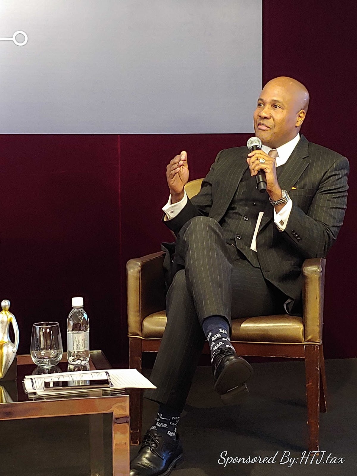 Man in pinstripe suit speaking into a microphone while seated.