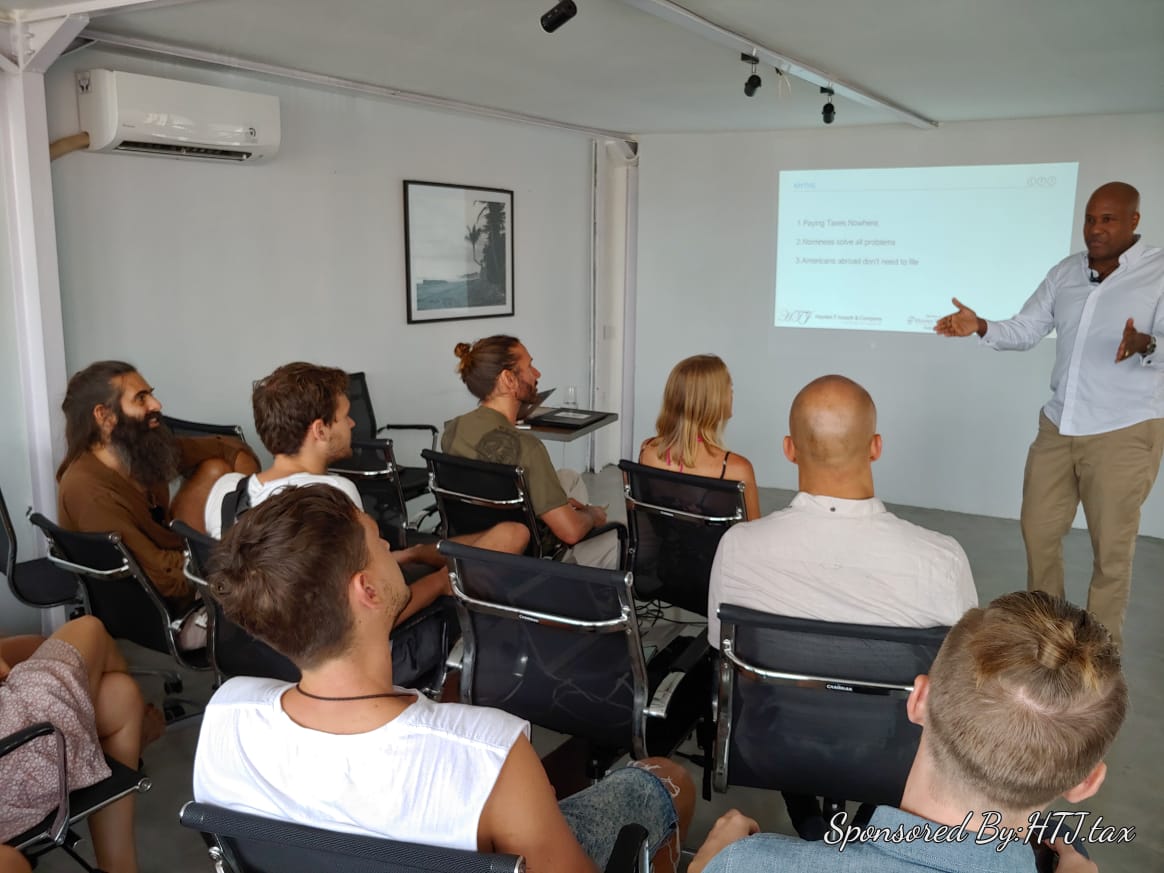 Speaker presents to an audience in front of a projector screen with bullet points.