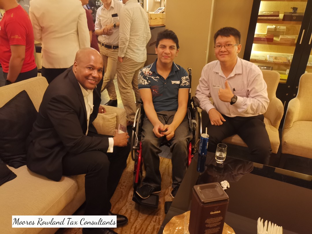 Three men posing for a photo, one in a wheelchair. Moores Rowland Tax Consultants text at bottom.