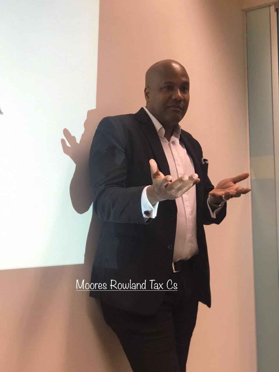 Man in suit gesturing with hands, Moores Rowland Tax Cs watermark.