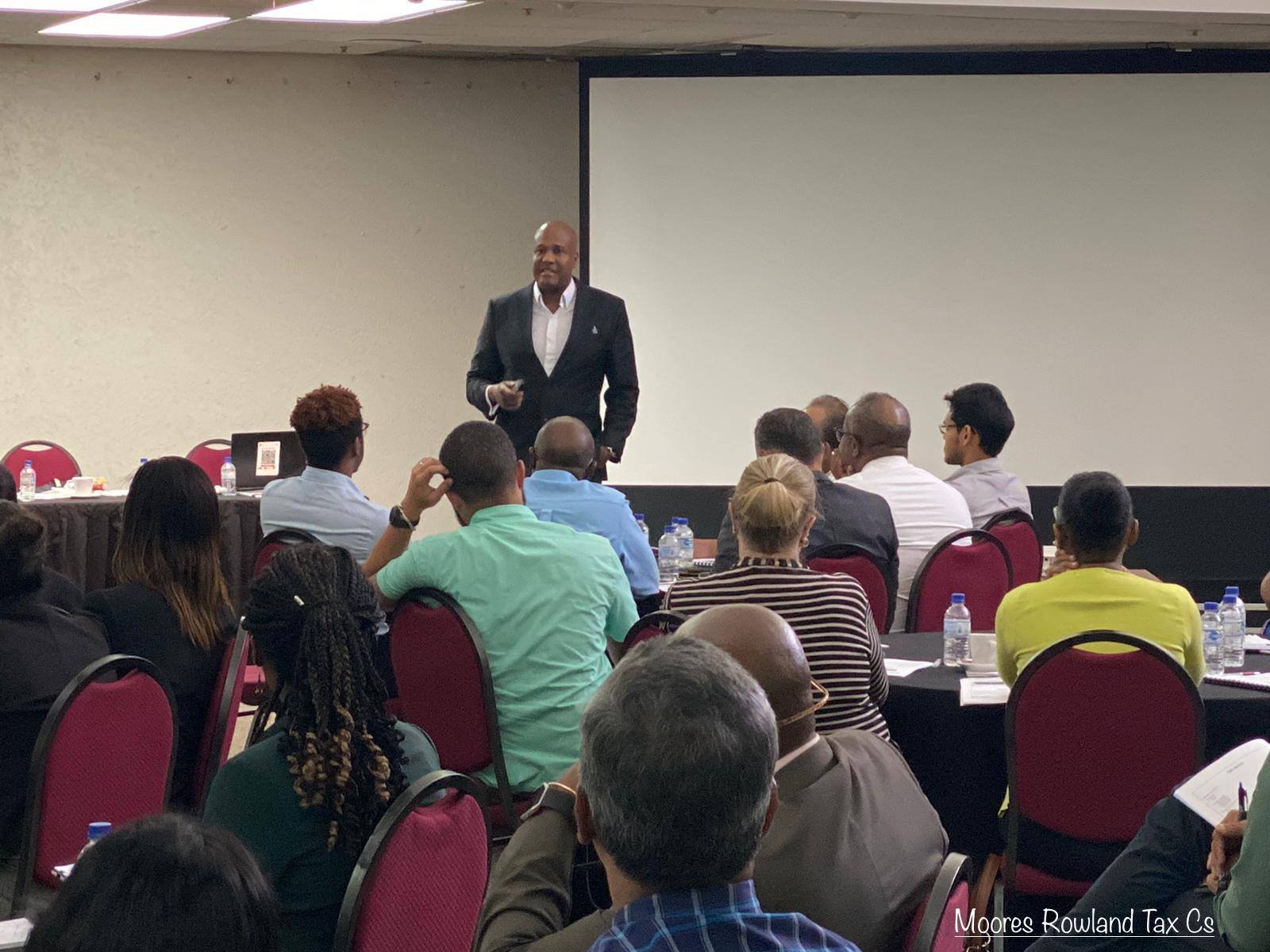 Man in suit presents to an audience seated at tables in a conference room.