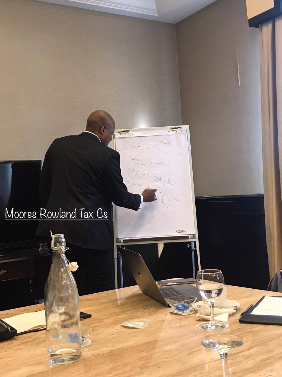 Man in suit writing on whiteboard with text Moores Rowland Tax Cs overlaid.