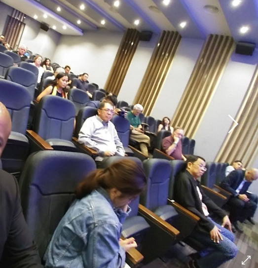 Audience seated in a lecture hall with blue chairs and wood paneling.