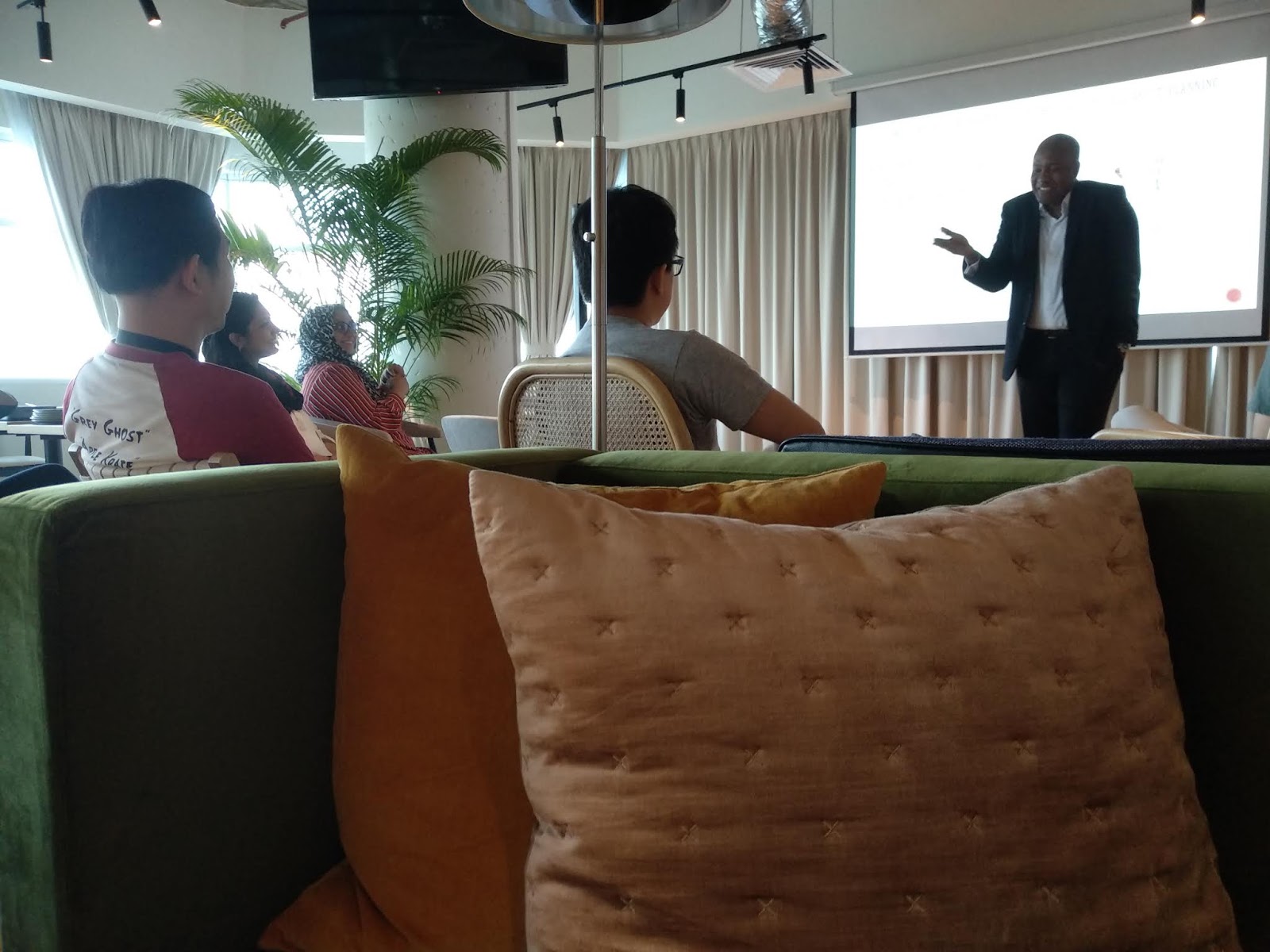 Man in suit presenting to an audience in a modern room with plants and couches.