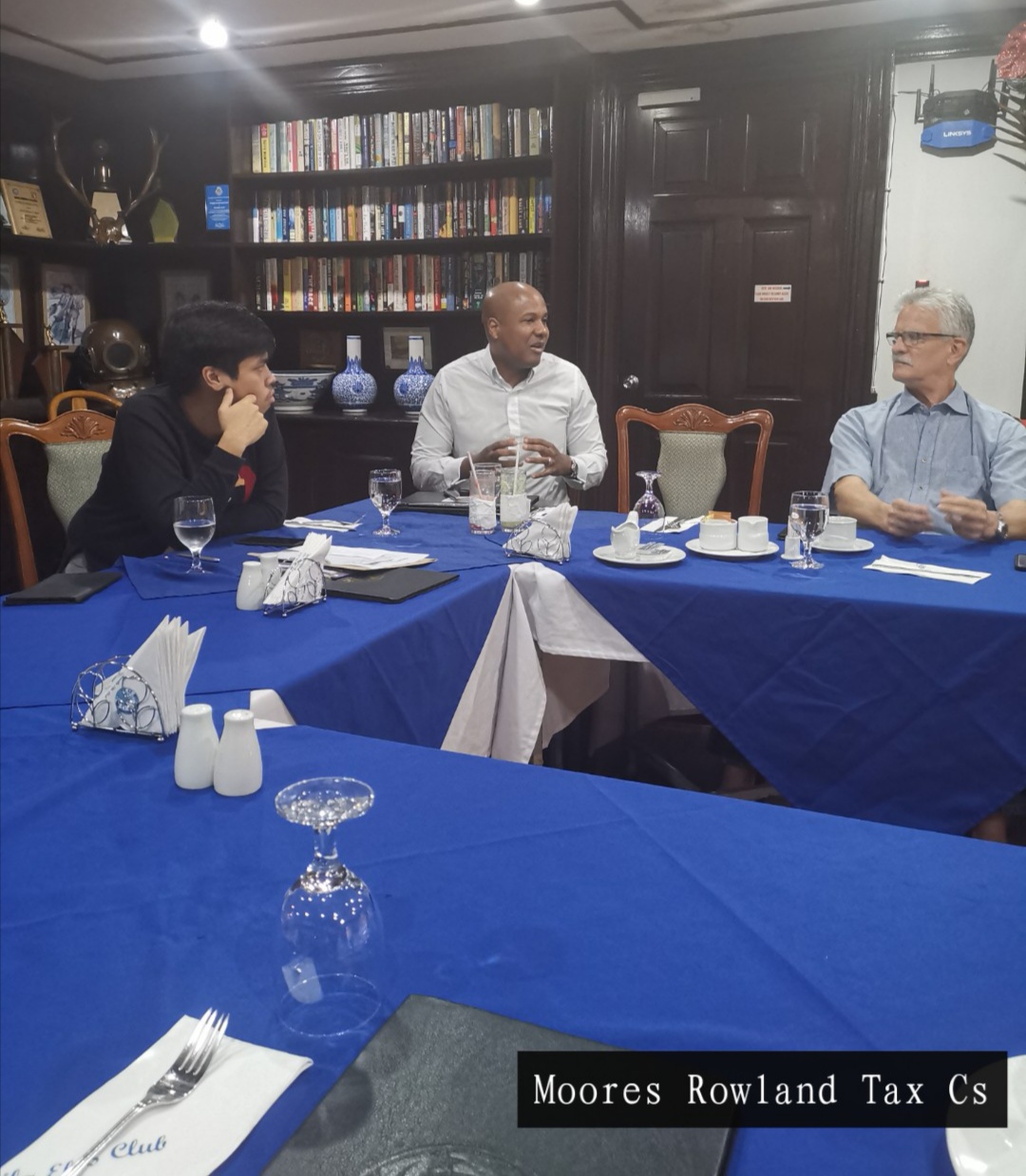 Three men sit at a table covered in blue tablecloths. Bookshelves are behind them.