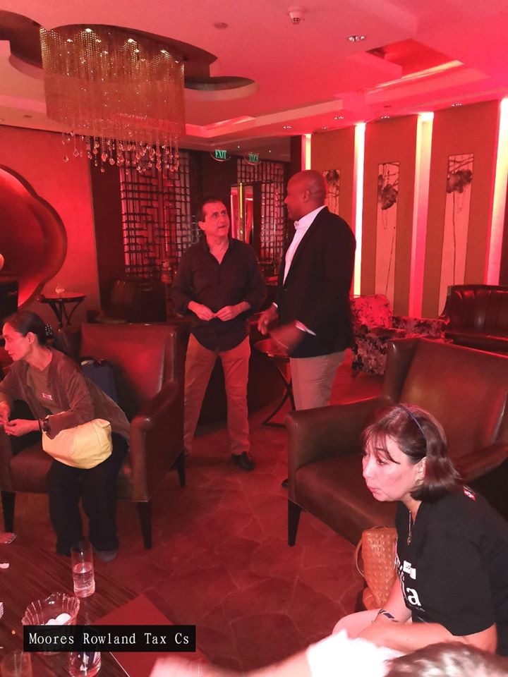 Moores Rowland Tax Cs logo on a red-lit lounge with people sitting and standing.