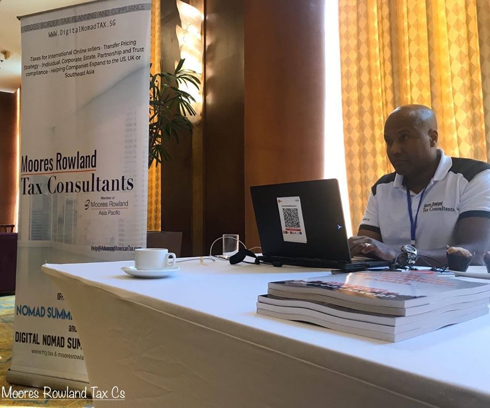 Man at laptop with Moores Rowland Tax Consultants banner and books on table.
