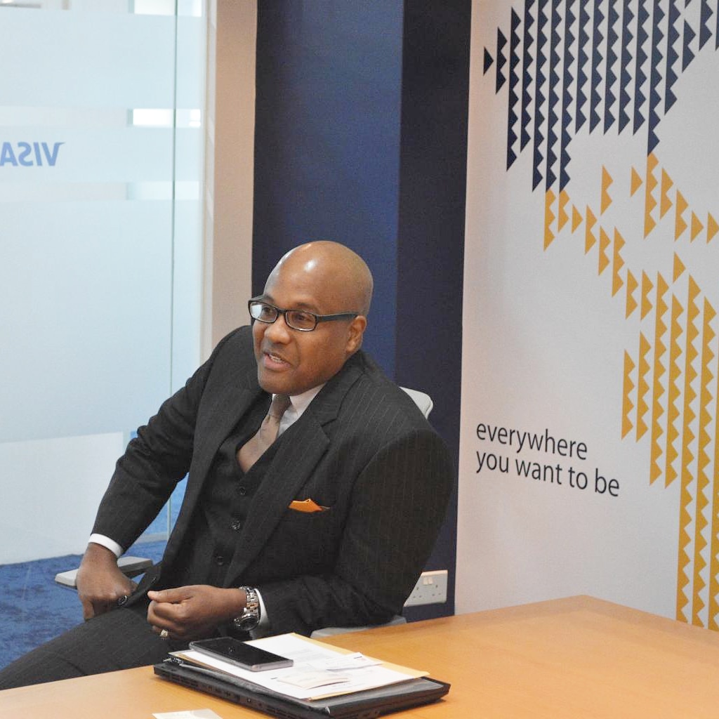 Man in suit and glasses sitting at desk with Visa branding in background.