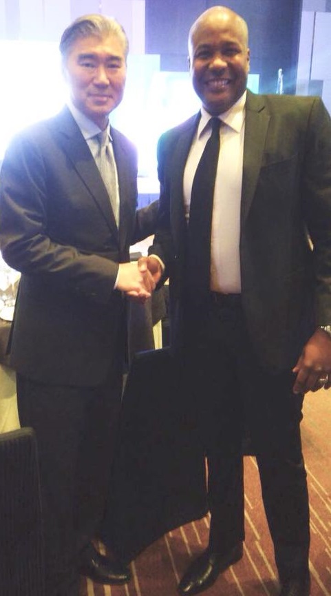 Two men in suits shake hands. One man is Asian with graying hair, the other is Black.