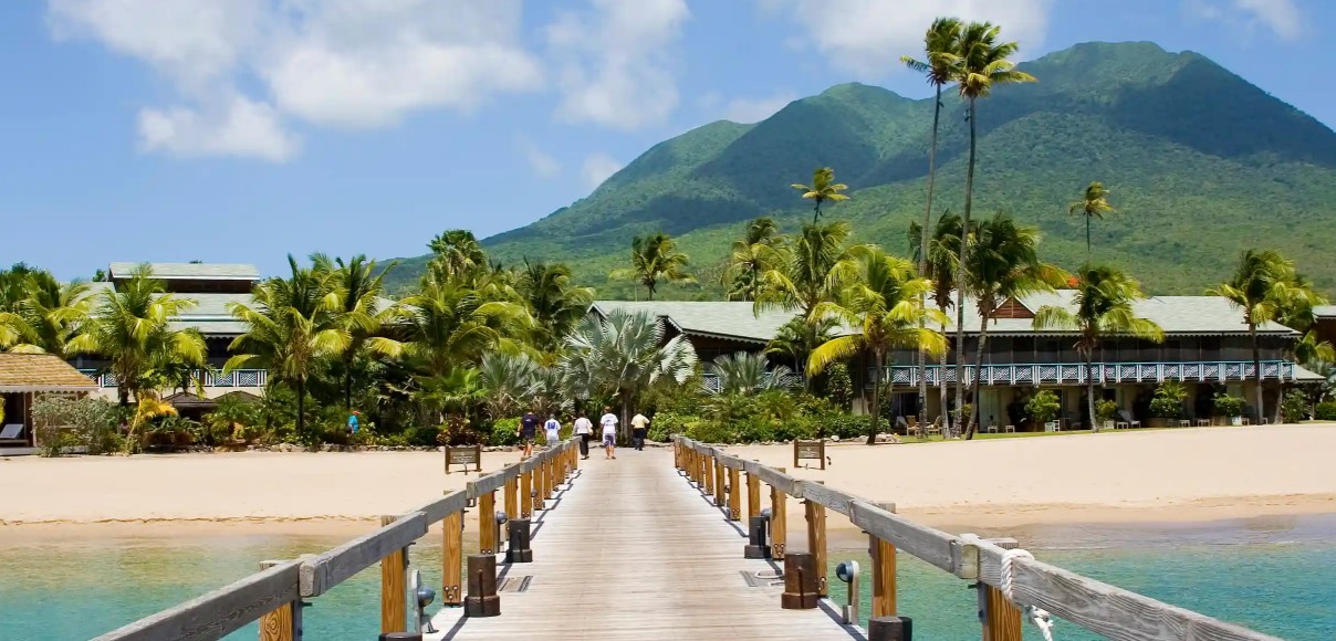 Wooden pier leading to a resort on a sandy beach with palm trees and a mountain.