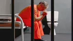 Man in orange jumpsuit sitting on a jail cell bed, head in hands.