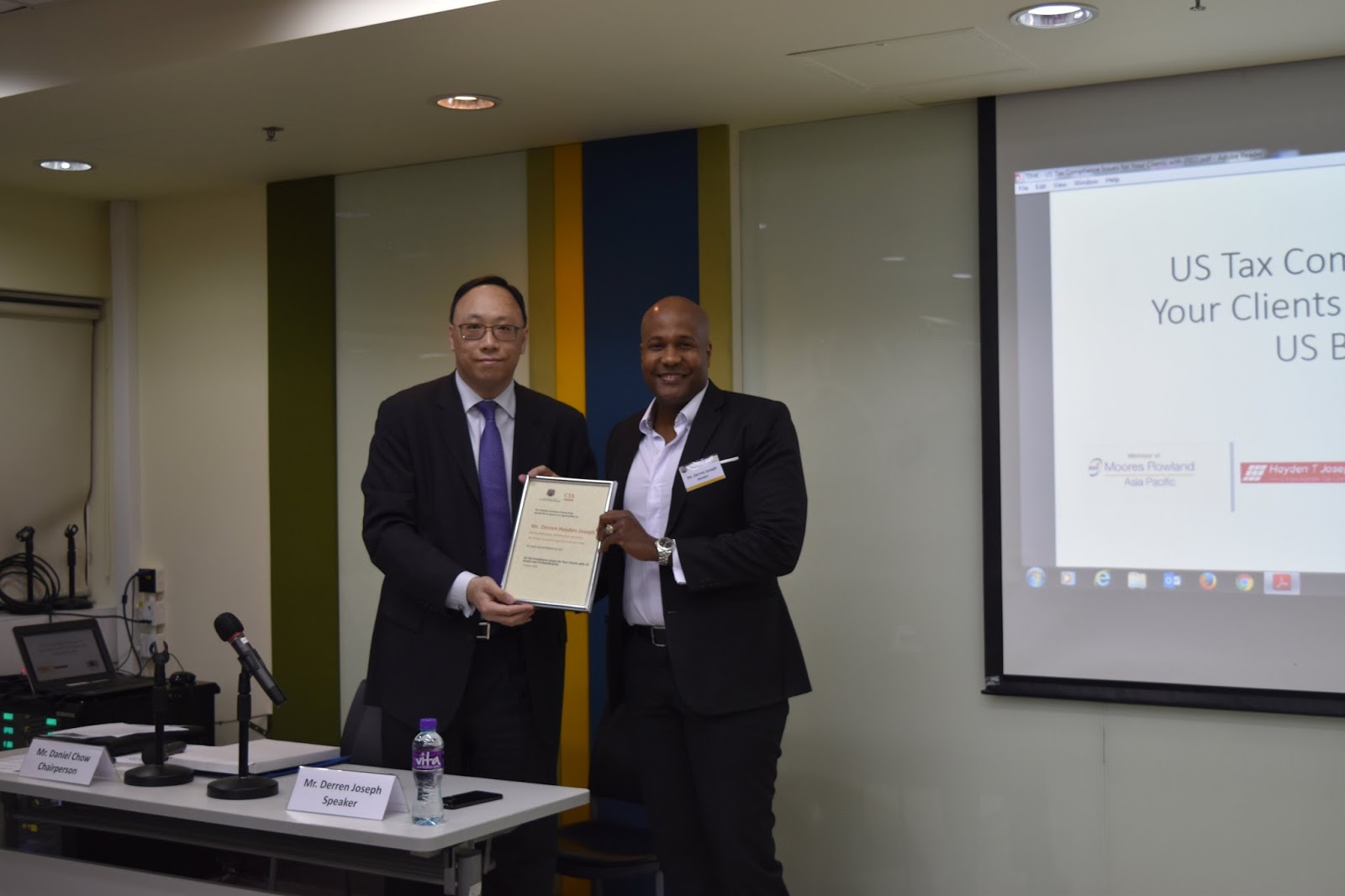 Two men holding a certificate, with a presentation screen behind them.