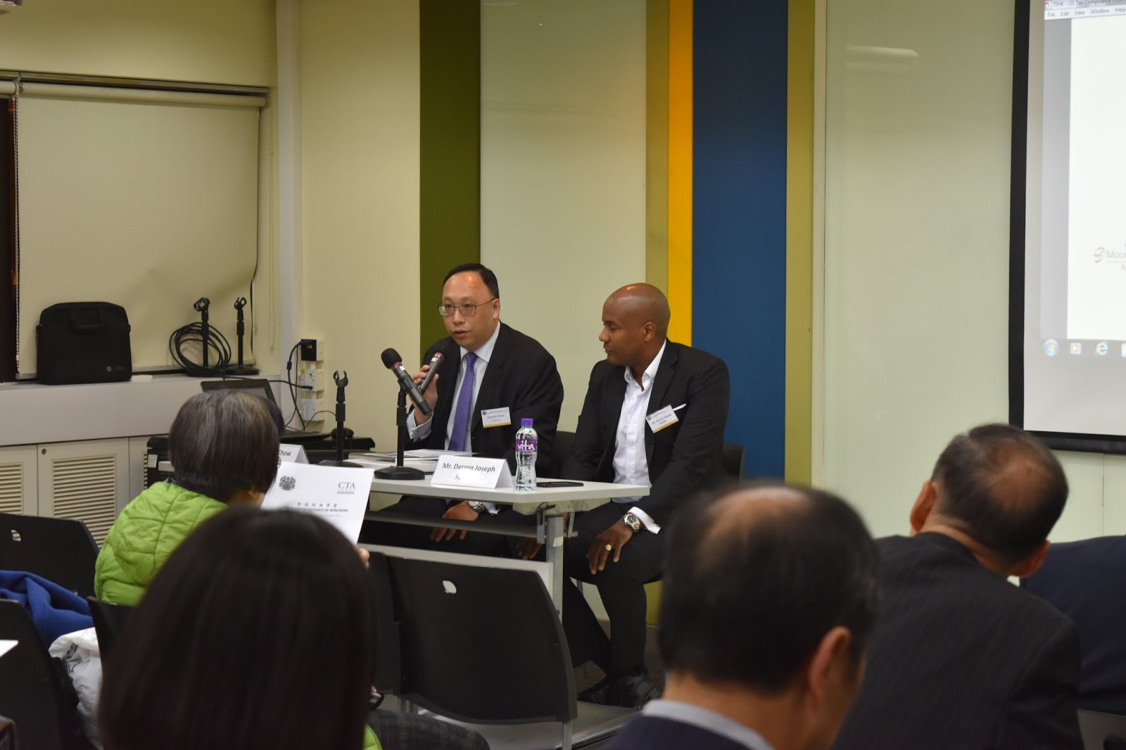 Two men speak at a panel table in front of an audience. One man holds a microphone.