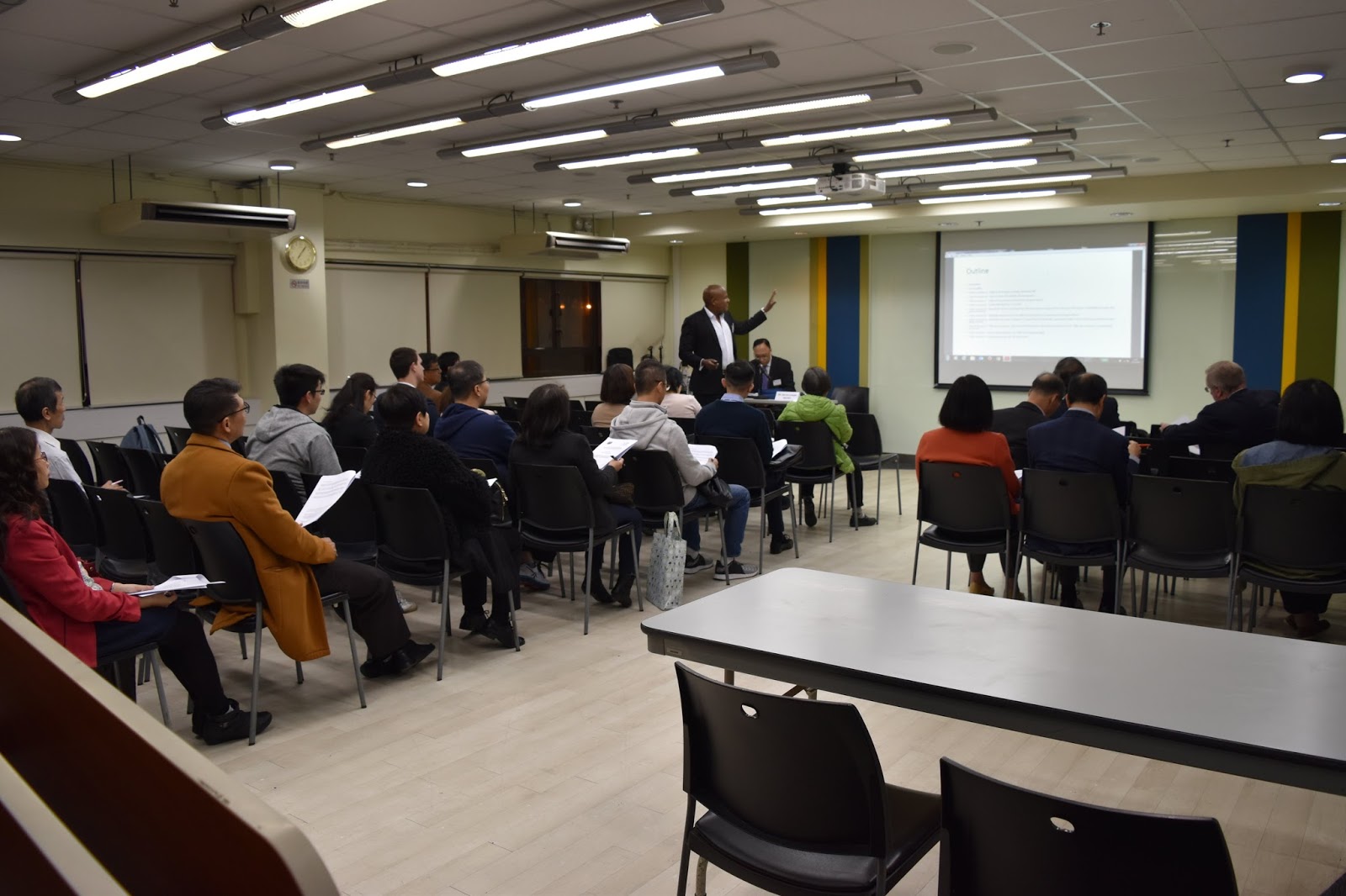 Speaker presenting to an audience seated in a conference room with a projector screen.