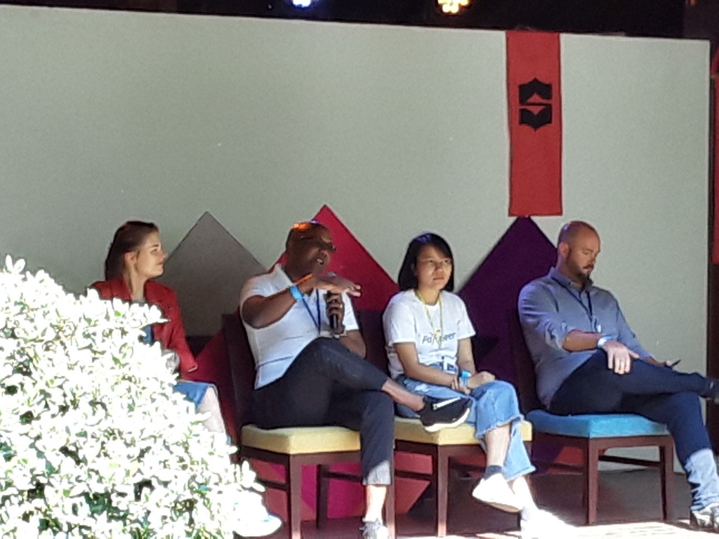 Four people sit on chairs on a stage, with one man speaking into a microphone.