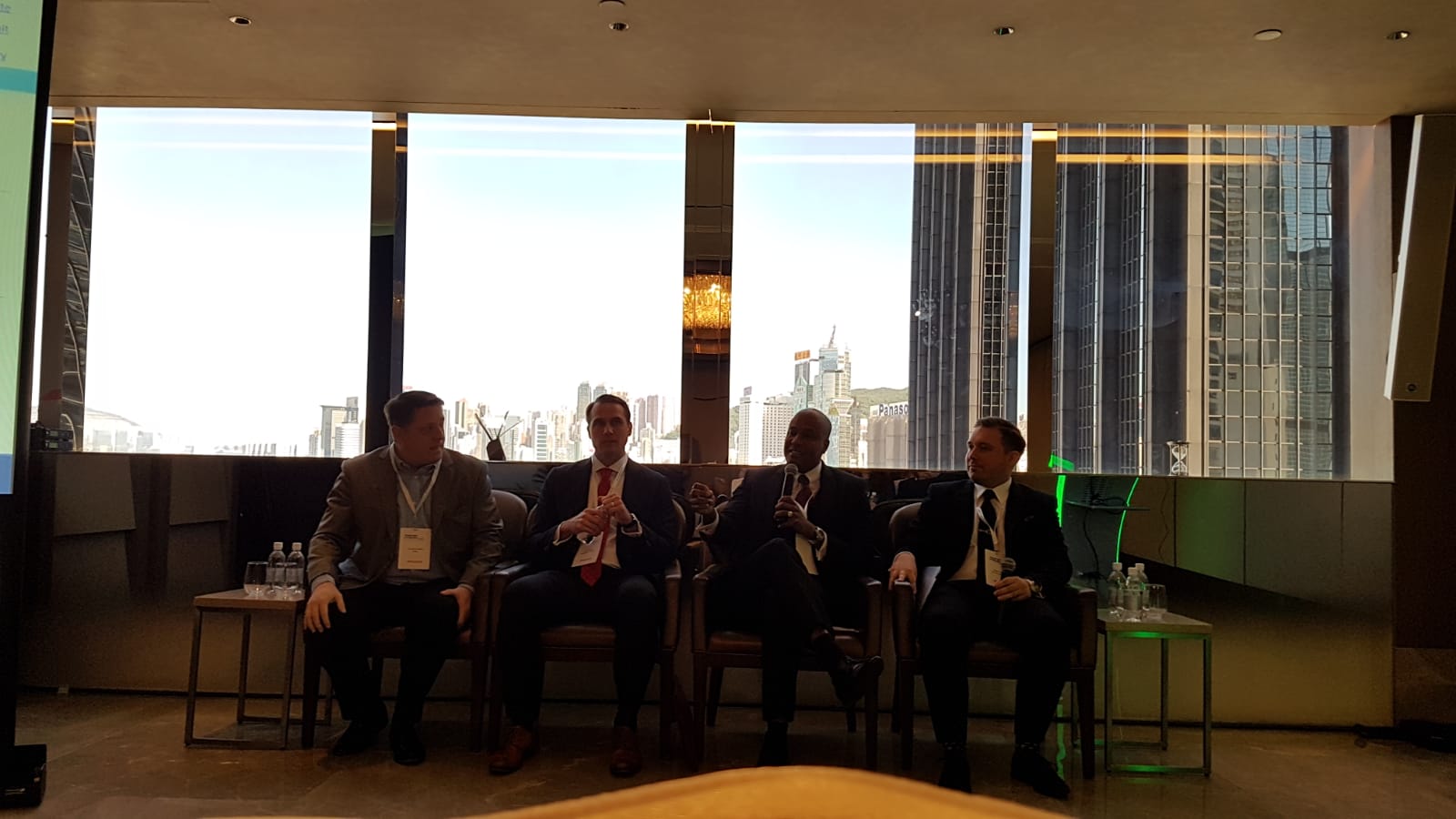 Four men in suits sit in chairs in front of a large window overlooking a city skyline.