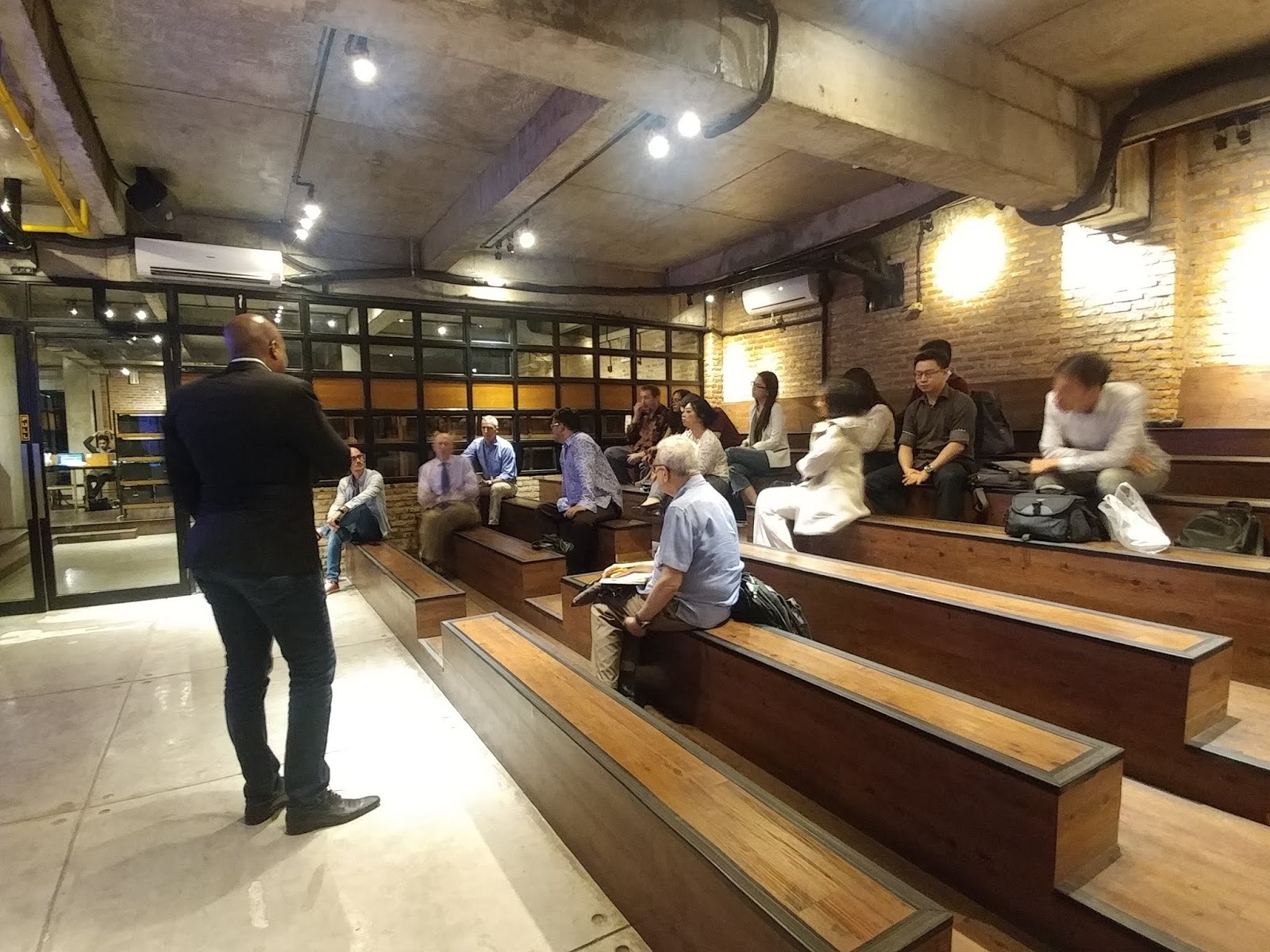 A man in a suit addresses an audience seated on tiered wooden benches in a modern room.